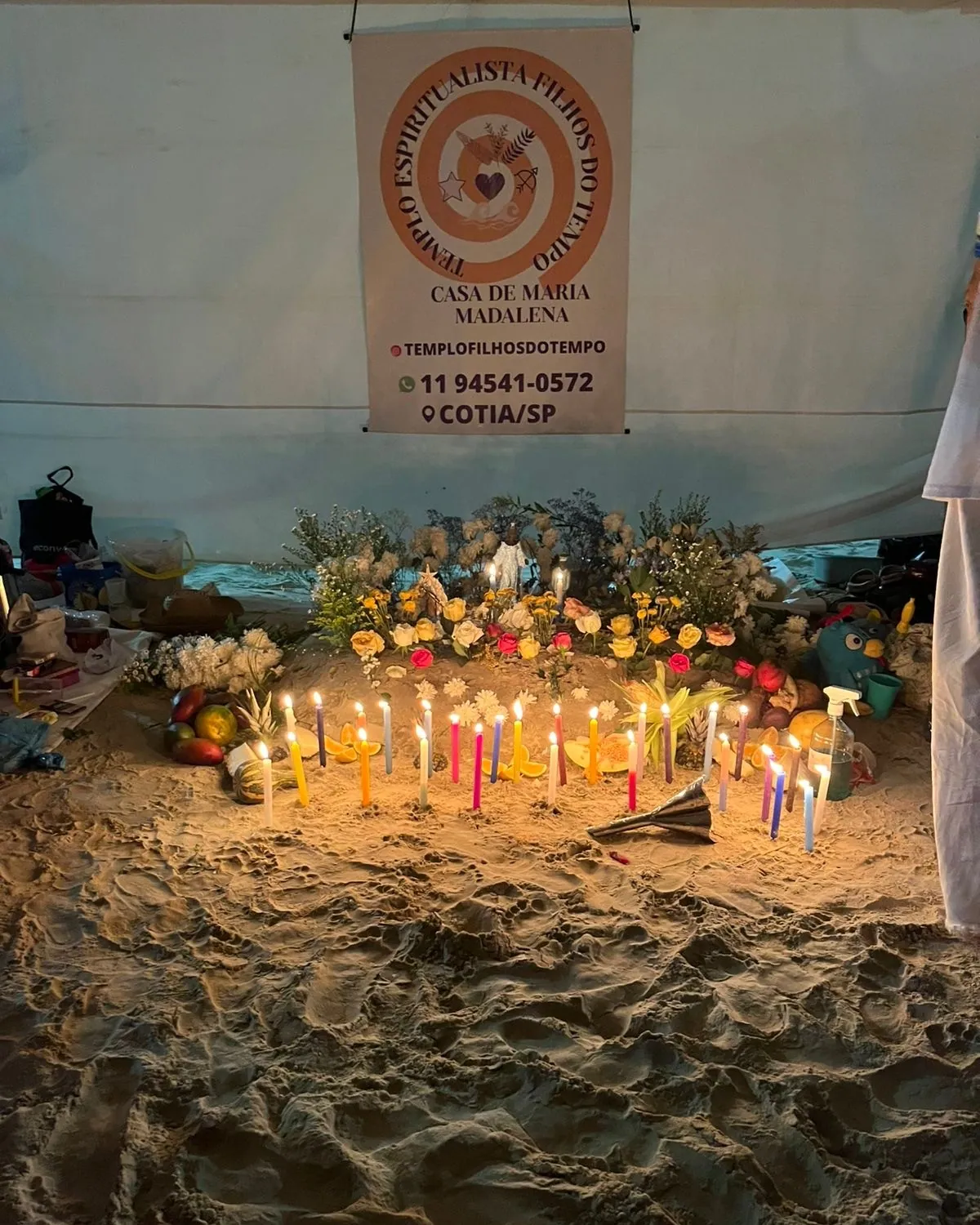Banner do templo com a espiral laranja sobre flores, frutas e velas coloridas acesas na areia da praia