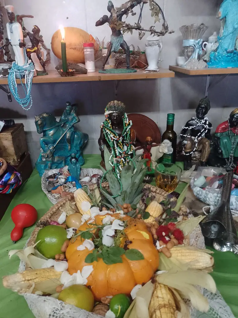 Oferenda em cesto com abóbora, milho, frutas e pétalas brancas diante das imagens do altar