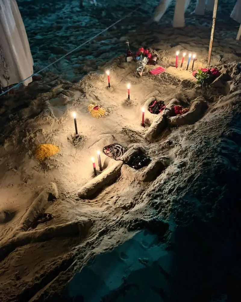 Velas vermelhas, pretas e azuis acesas na areia à noite, entre flores, guias e oferendas